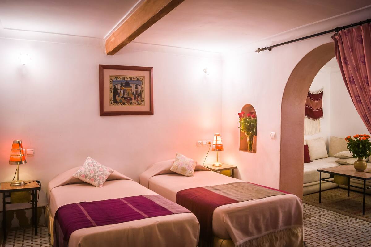 A hotel room with two beds, a small table with a lamp, a framed picture on the wall, and a view of the living area through an archway.