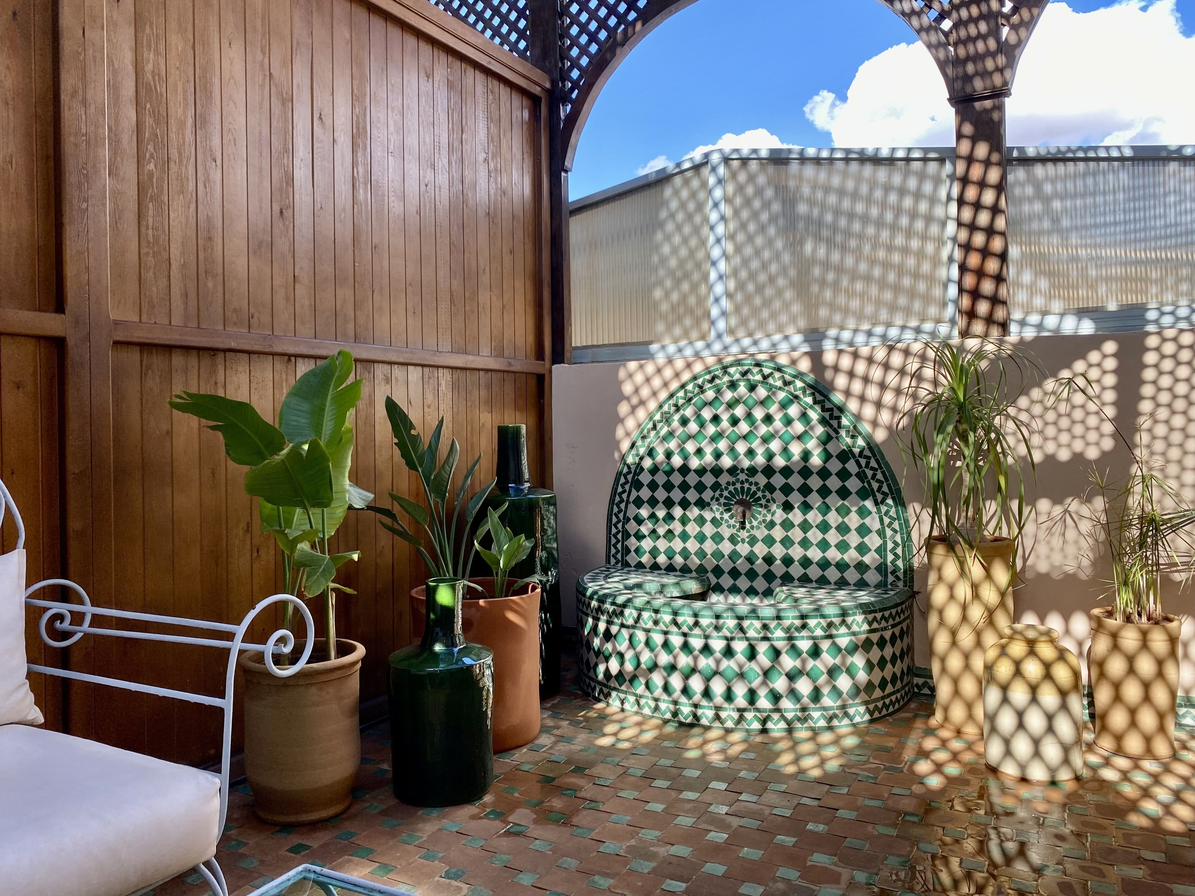 A sunlit patio with a wooden wall, a patterned outdoor seating area, and potted plants.