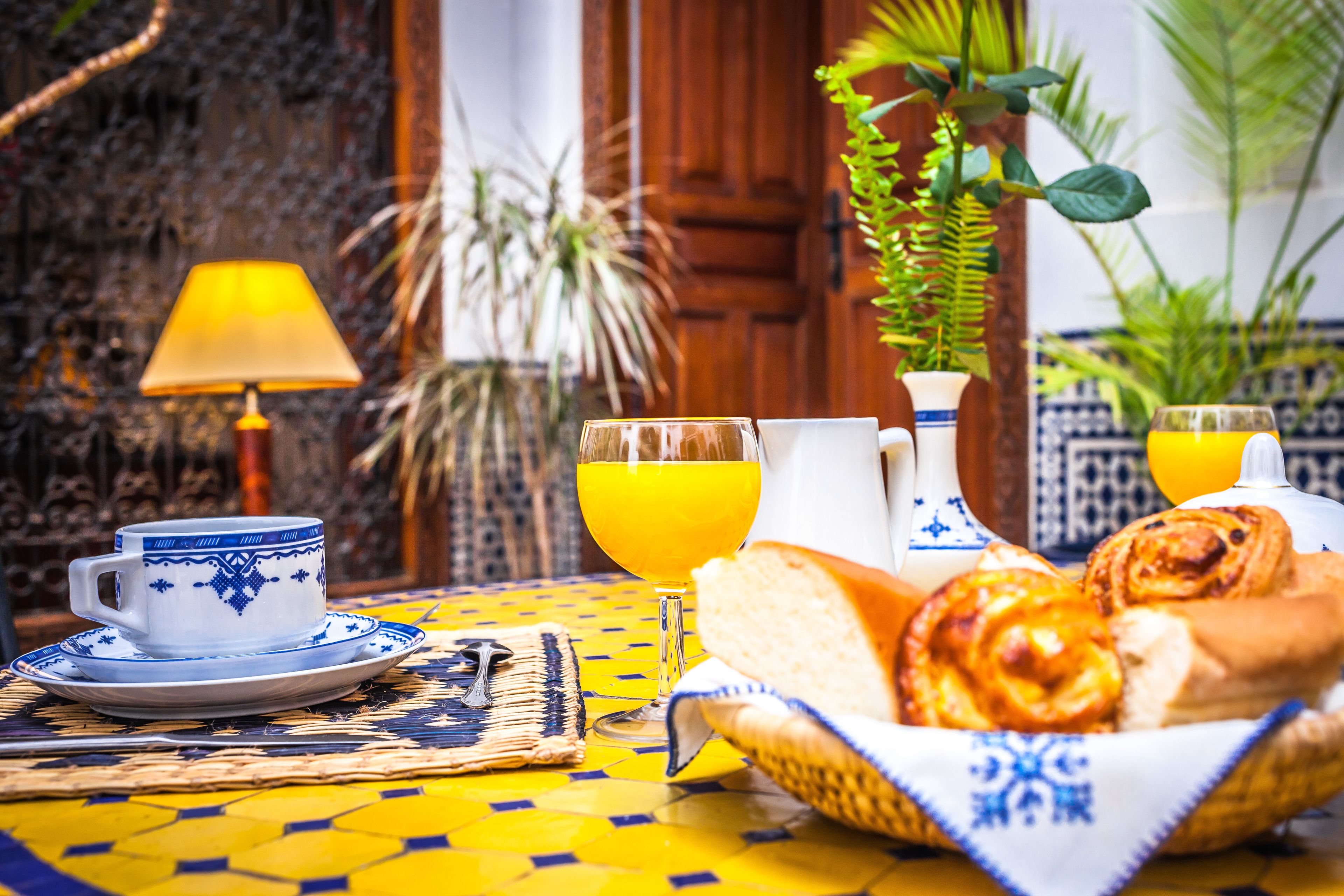 Une table de petit-déjeuner avec une tasse, un verre de jus d’orange, une carafe et un panier de viennoiseries.