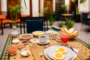 Mesa posta para o pequeno-almoço, com vários pratos, incluindo ovos, pão e bebidas, sobre uma toalha de mesa estampada.