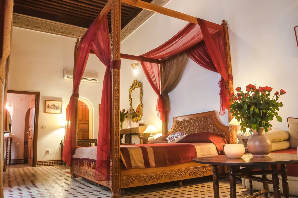 A four-poster canopy bed with red drapes, a wooden headboard, and a vase of flowers on a bedside table.