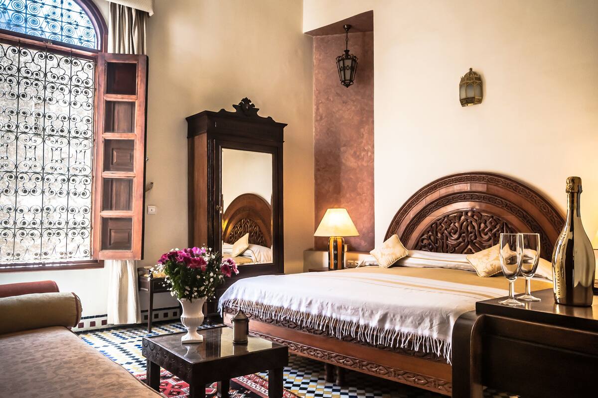 A hotel room with a large bed, a wooden headboard, a nightstand with a lamp, a vase with flowers, and a window with decorative iron bars.
