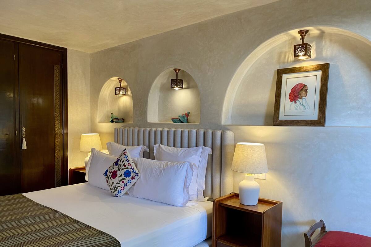 A hotel room with a large bed, two bedside tables, a nightstand, a chair, a wardrobe, and a framed picture on the wall.