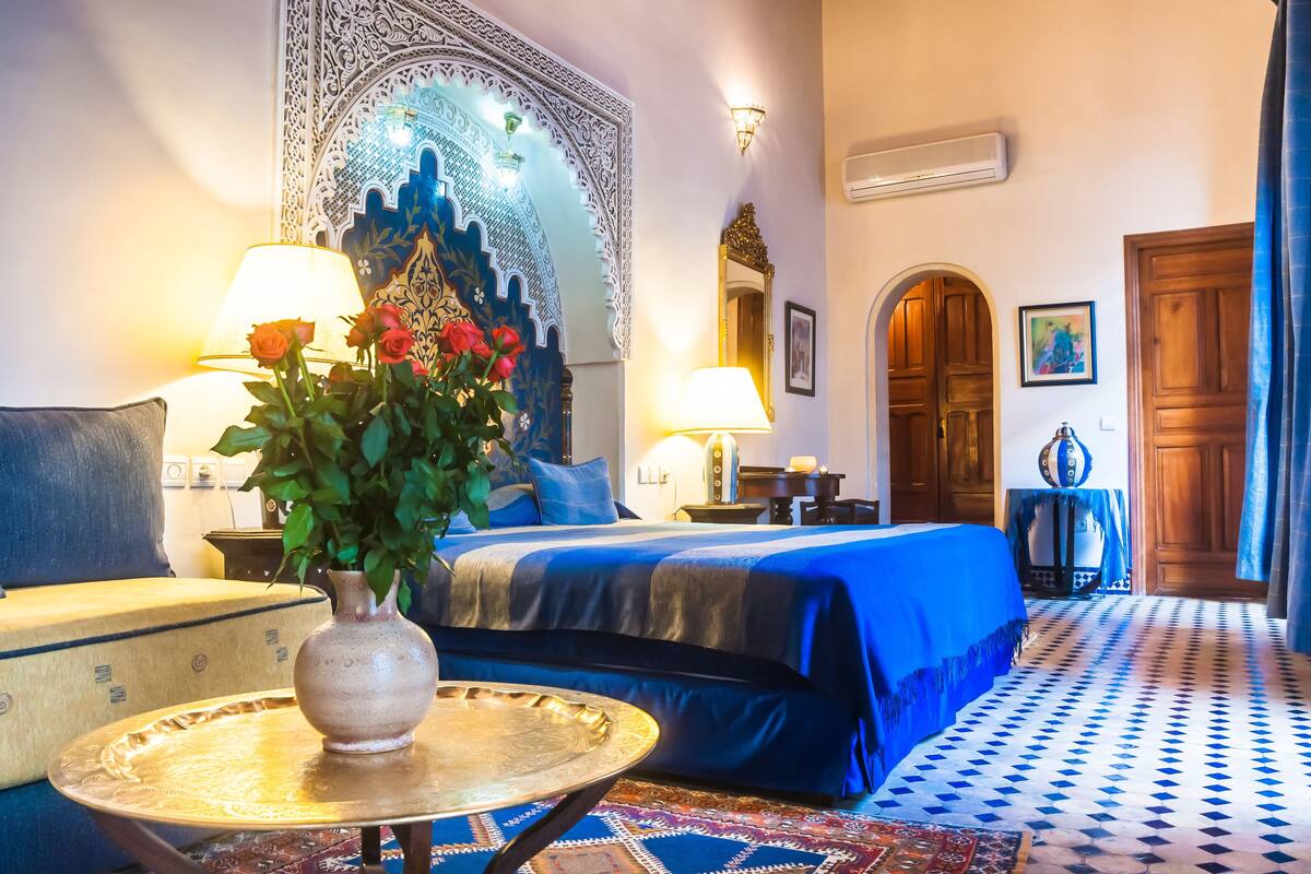 A hotel room with a bed, a sitting area with a table and chairs, a decorative wall hanging, and a patterned floor.