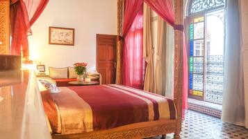 A traditional room with a bed, a small table with a vase, and a view of a courtyard through a window with decorative ironwork.