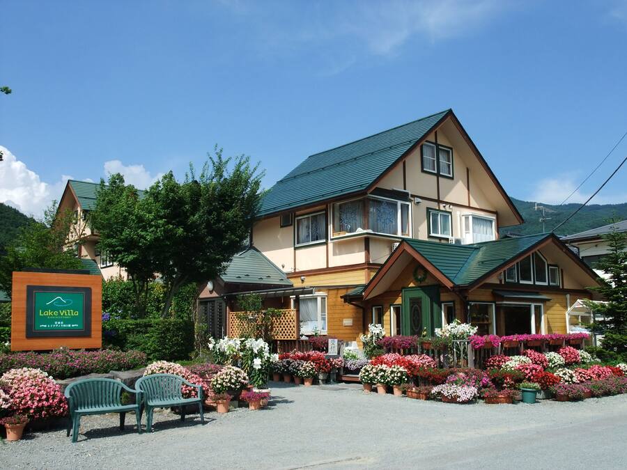 A two-story building with a green roof, surrounded by a variety of potted flowers and a bench.