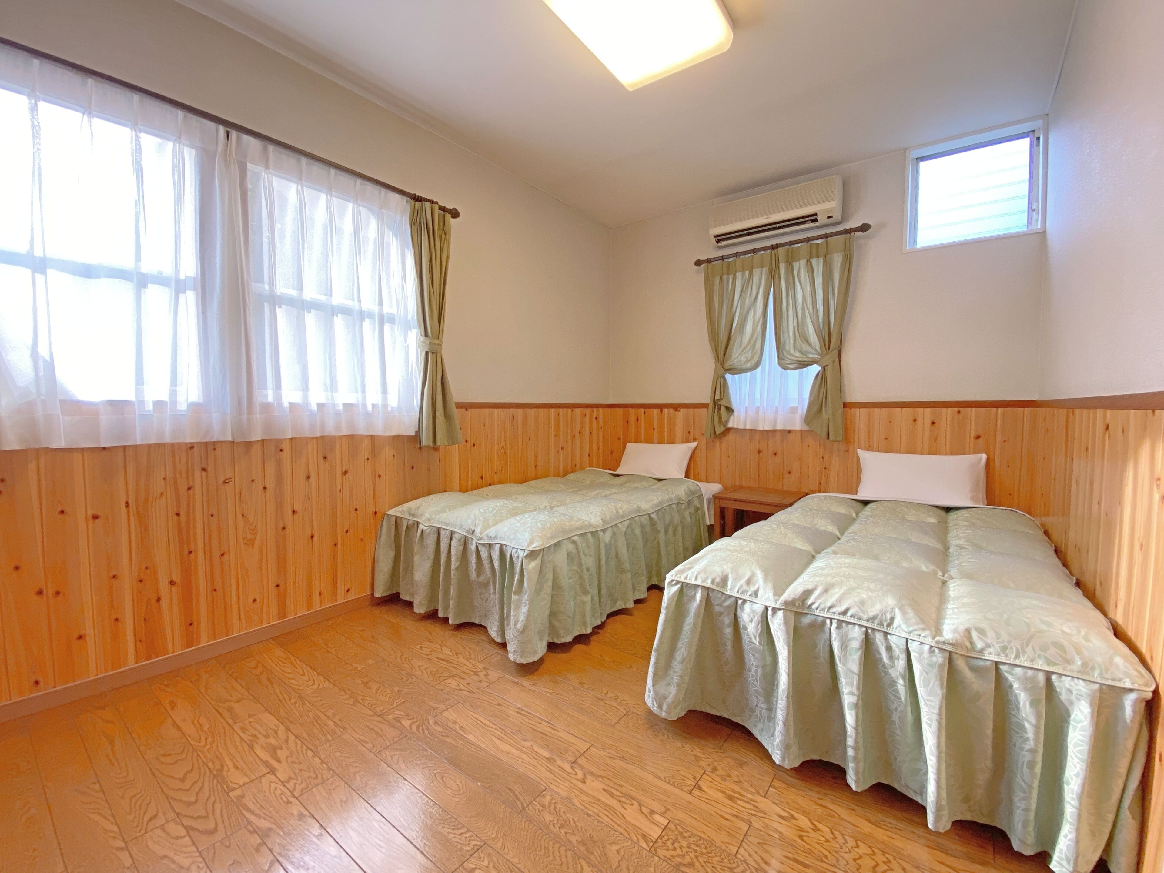 A room with two beds, wooden walls, a ceiling light, an air conditioning unit, and windows with curtains.
