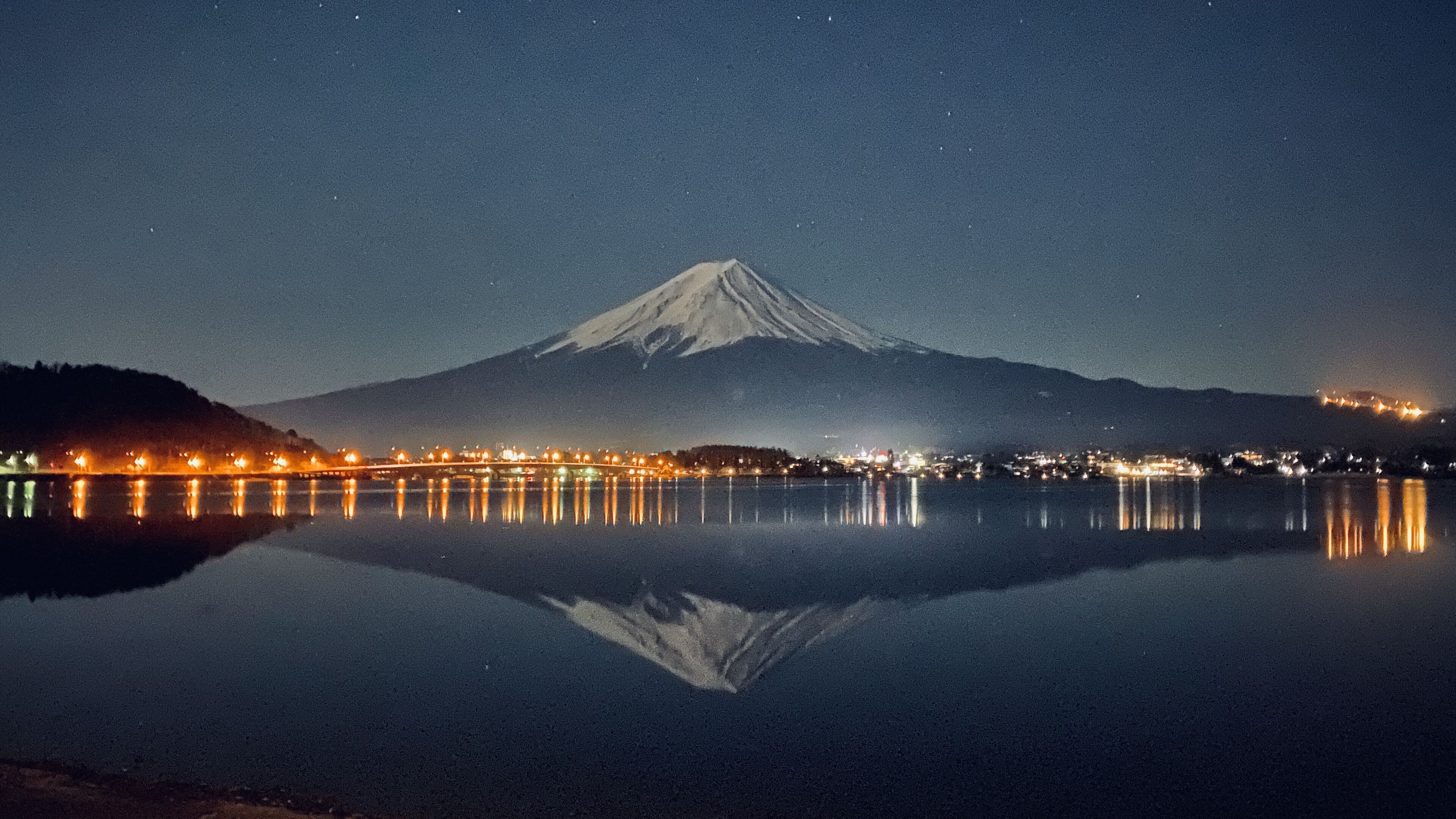 A nighttime view of a mountain with a snow-capped peak, reflecting on a calm body of water, with city lights along the water's edge.