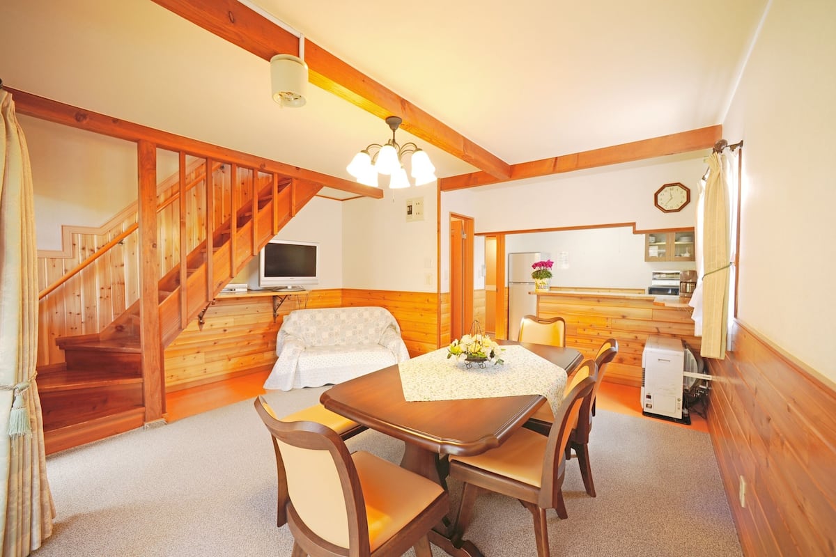 Une pièce chaleureuse avec un escalier en bois, un canapé, une table à manger avec des chaises, un petit coin cuisine et une horloge au mur.