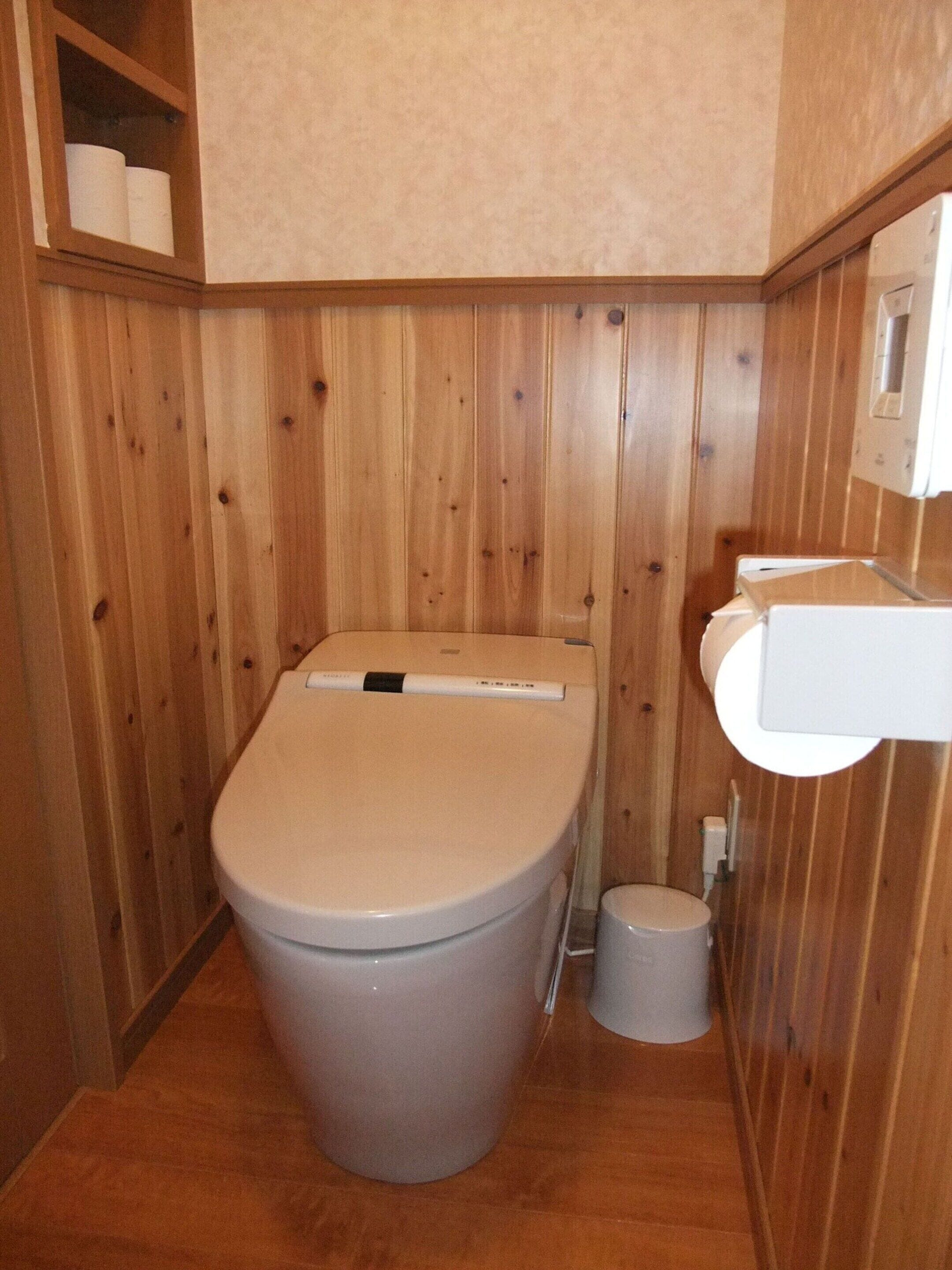 A small, wooden-paneled bathroom with a toilet, a bidet, and a shelf with toilet paper.