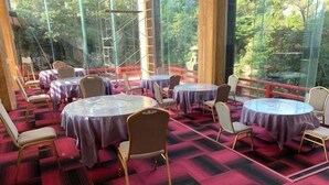Una habitación con mesas y sillas redondas, ventanas amplias con vistas a árboles y un suelo cubierto de alfombra roja.