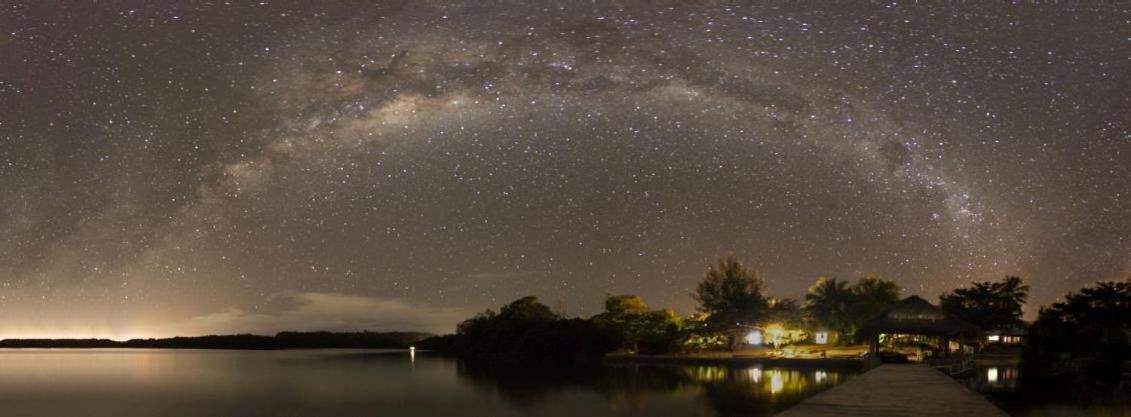A night sky with stars and the Milky Way over a calm lake, with a dock and buildings in the foreground.