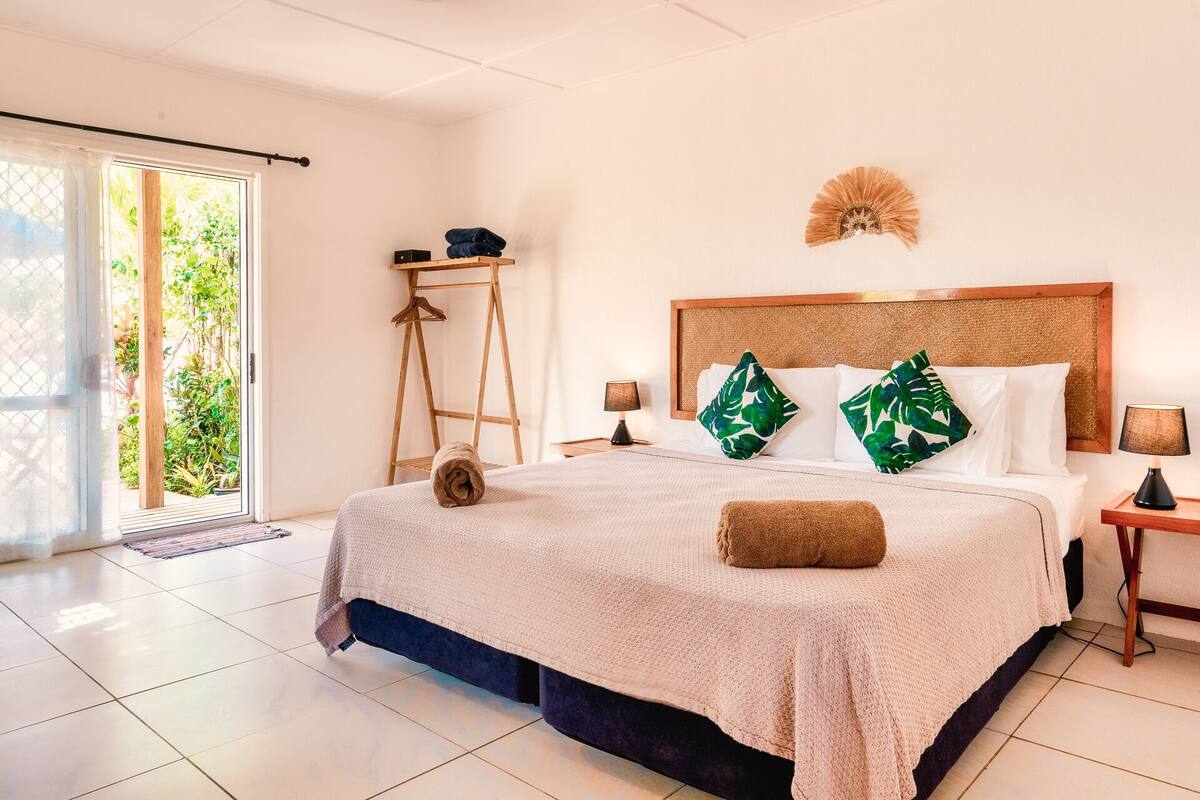 Un dormitorio con una cama grande, una escalera de madera, una mesita con una lámpara y vistas a la vegetación a través de una puerta corredera.
