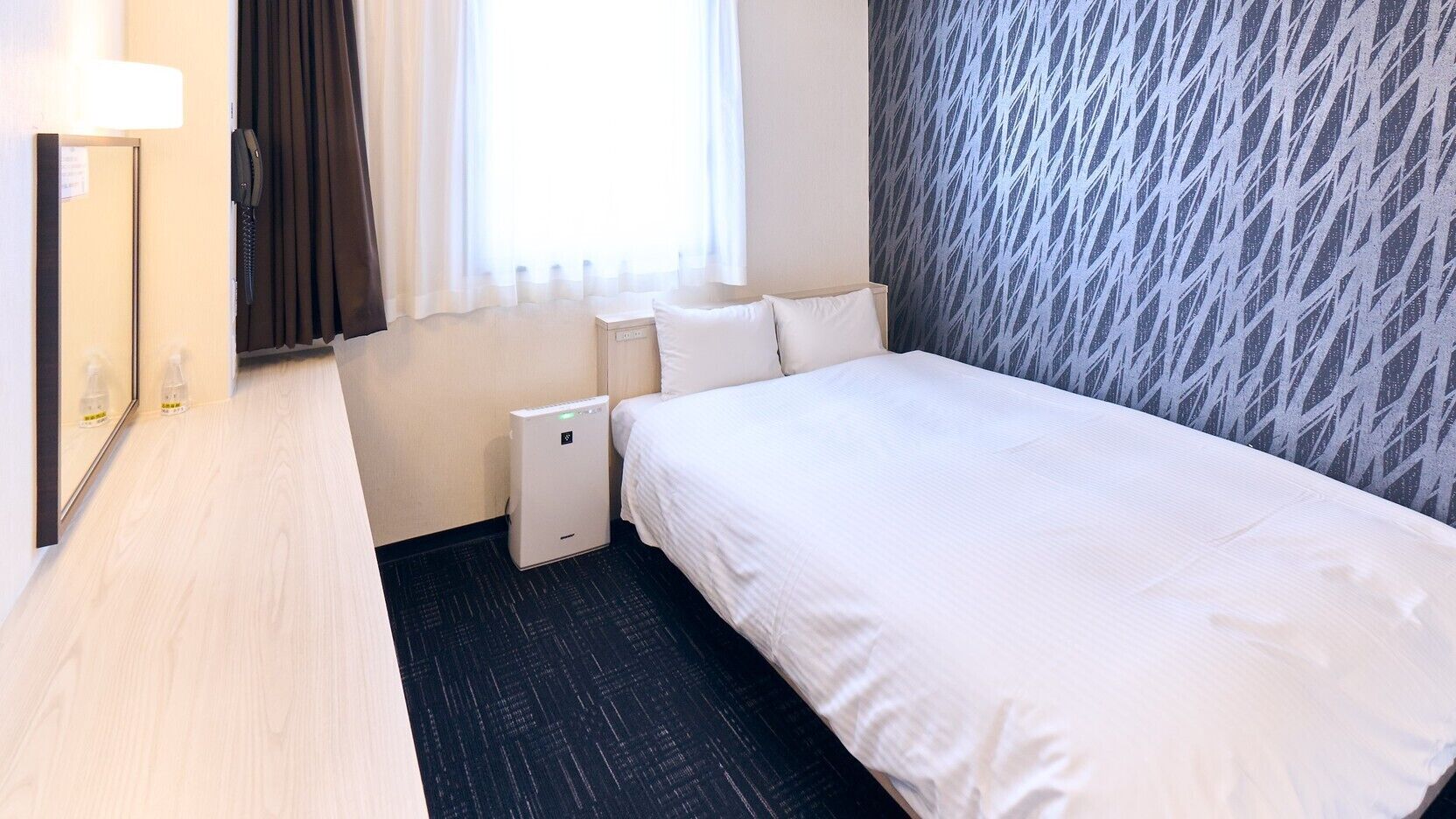 A hotel room with a bed, bedside table, lamp, and a small heater.