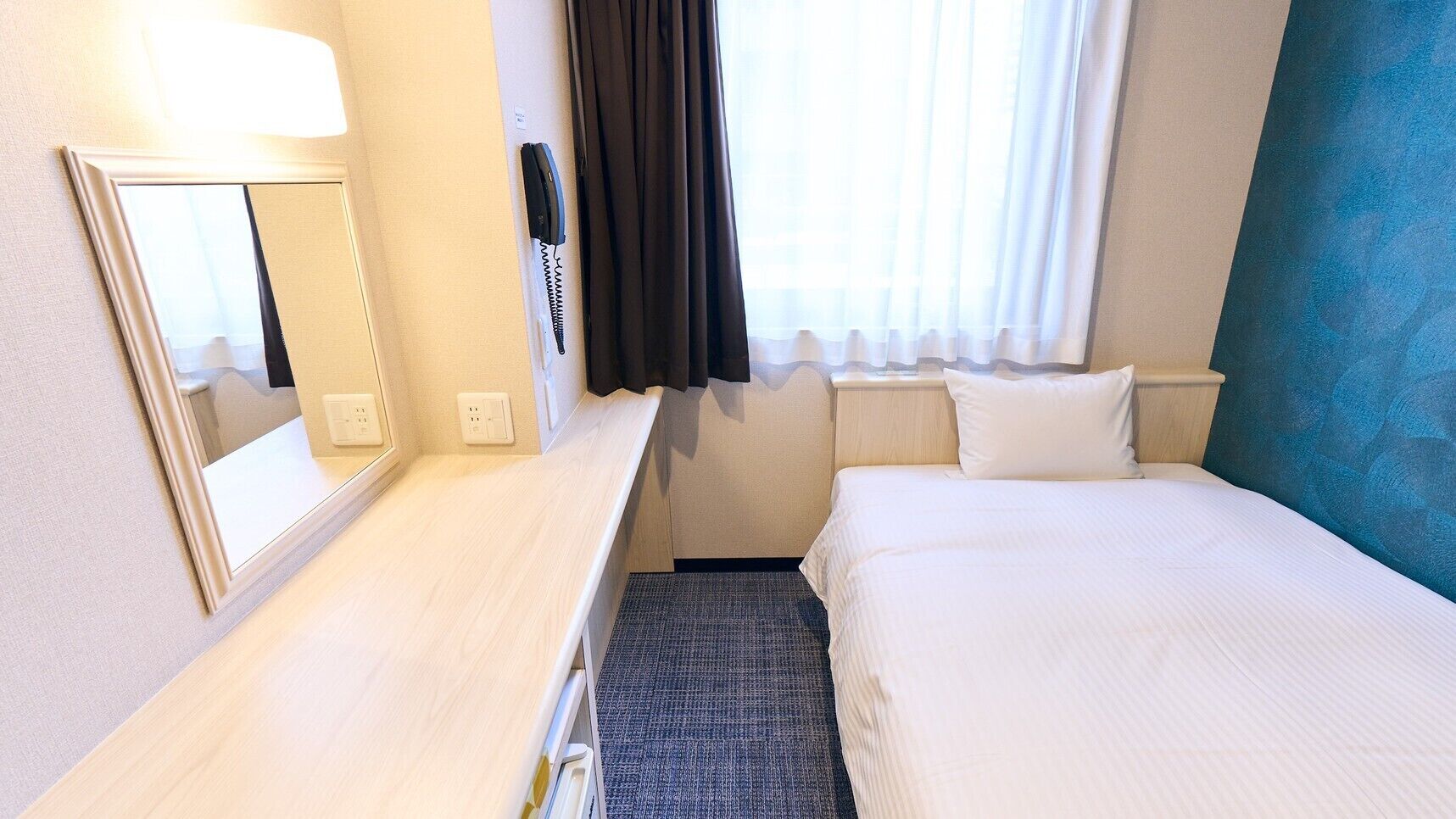 A hotel room with a bed, a desk with a mirror, a telephone, and a window with curtains.