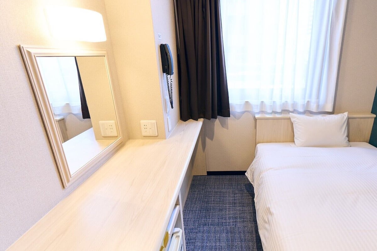 A hotel room with a bed, a desk with a mirror, a telephone, and a window with curtains.