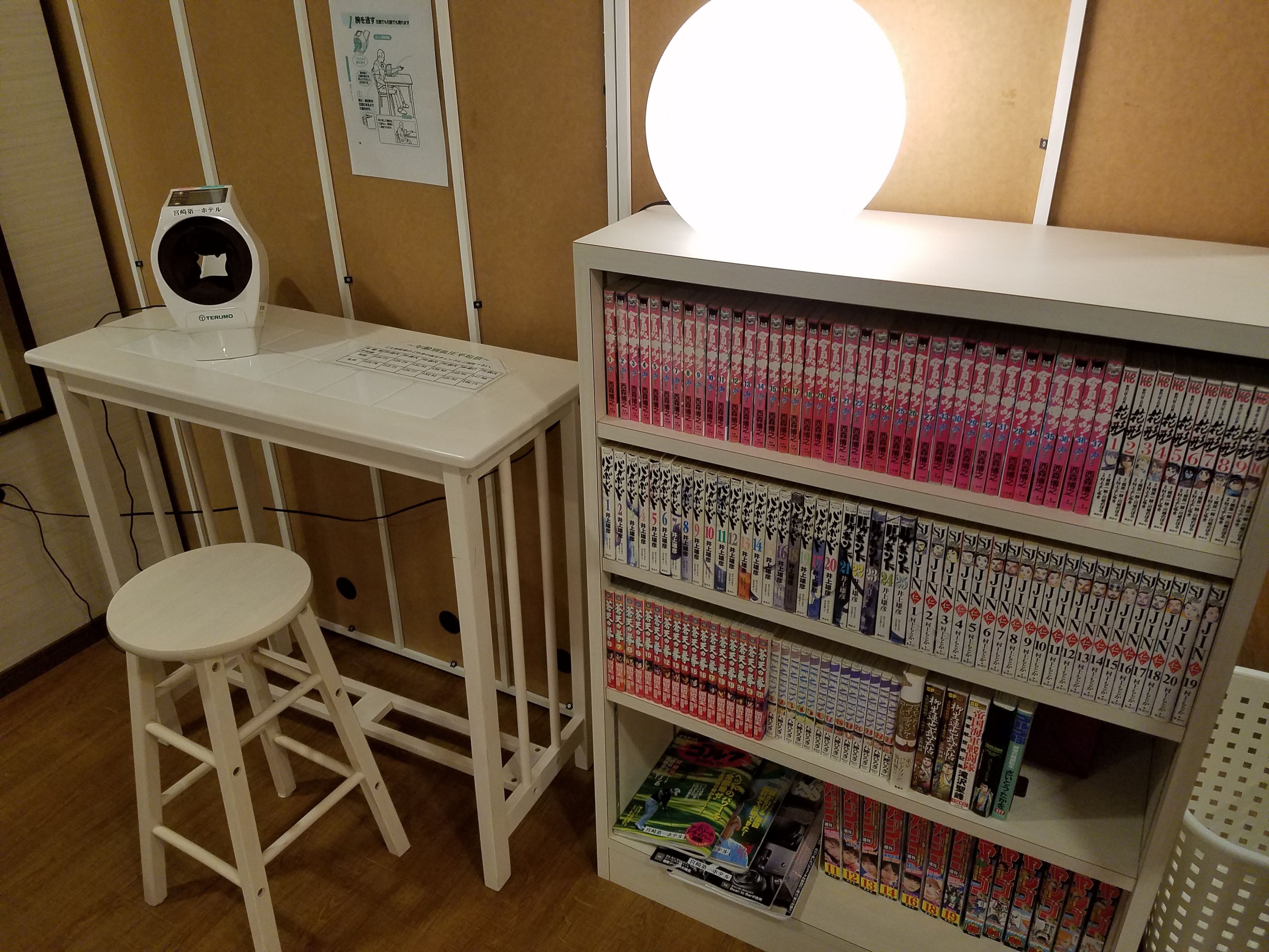 A room with a white desk, a stool, a shelf with books, and a round light fixture.