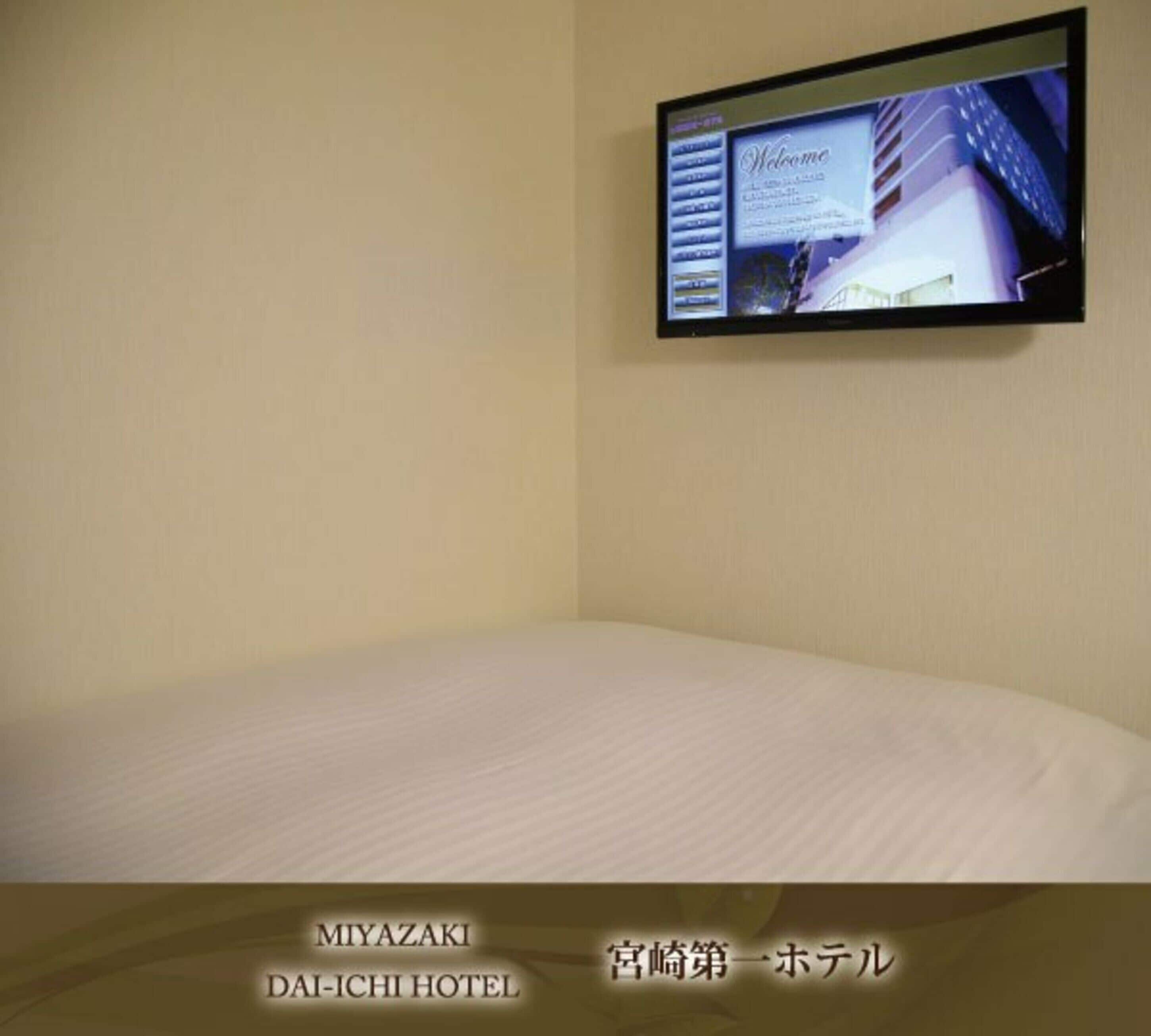 A hotel room with a bed, a television mounted on the wall displaying a welcome screen, and the hotel's name and location at the bottom.