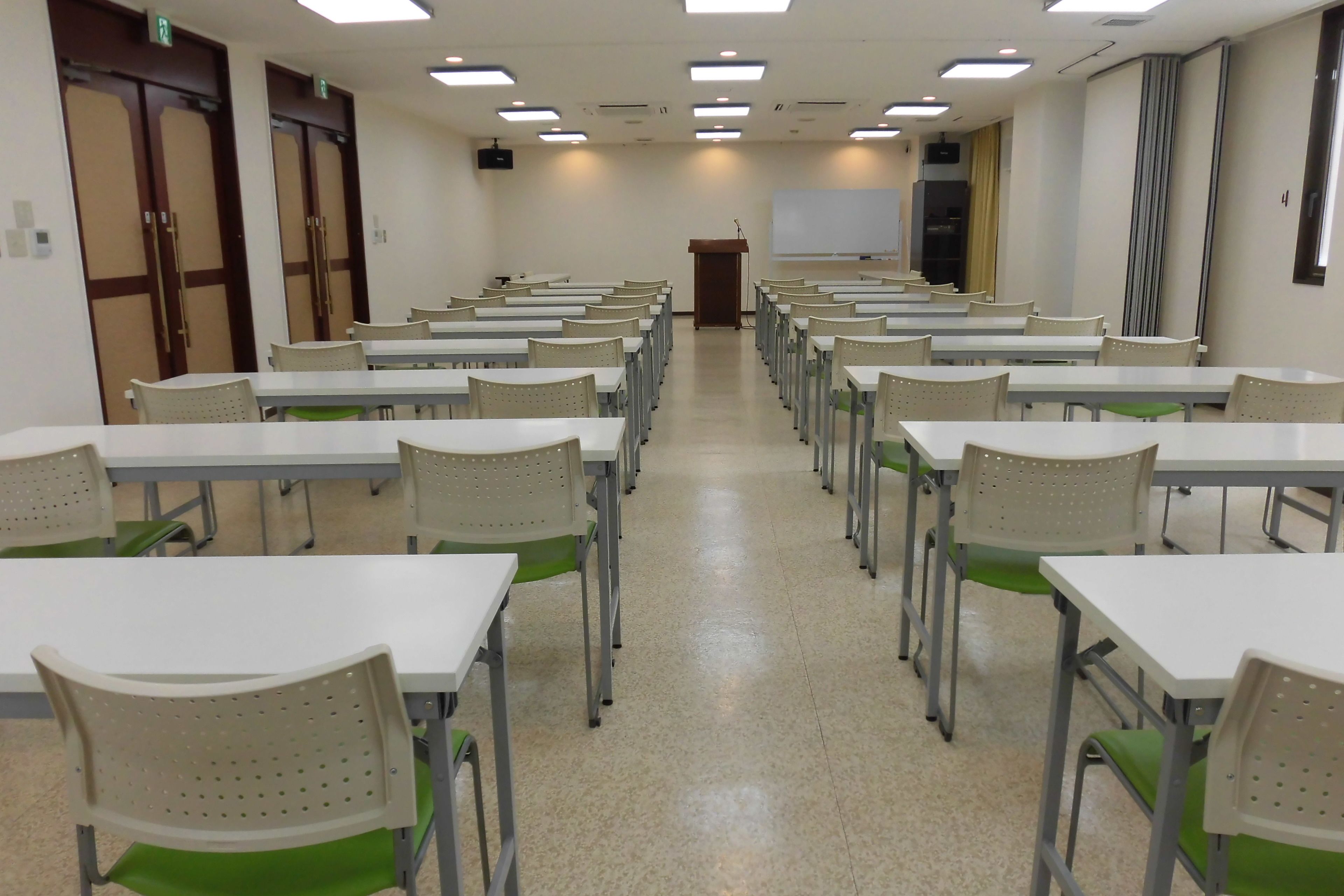 Un aula con filas de mesas y sillas, un atril al frente y una pizarra en la pared.