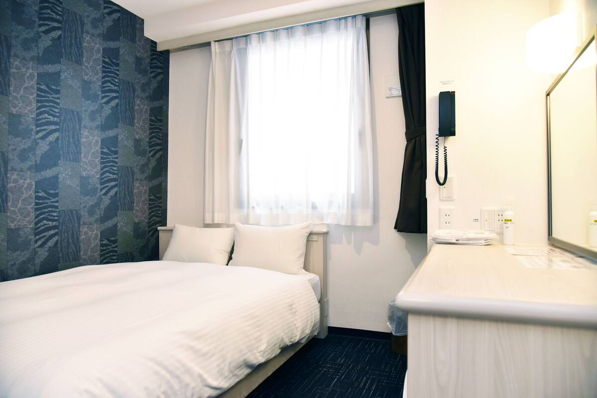 A hotel room with a bed, a bedside table, a telephone, and a window with curtains.