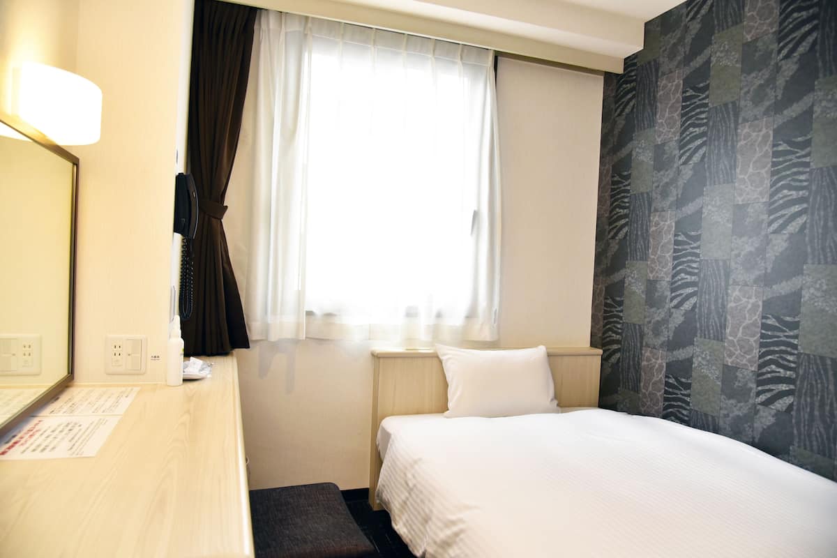 A hotel room with a bed, a bedside table, a mirror, a window with curtains, and a patterned wall.