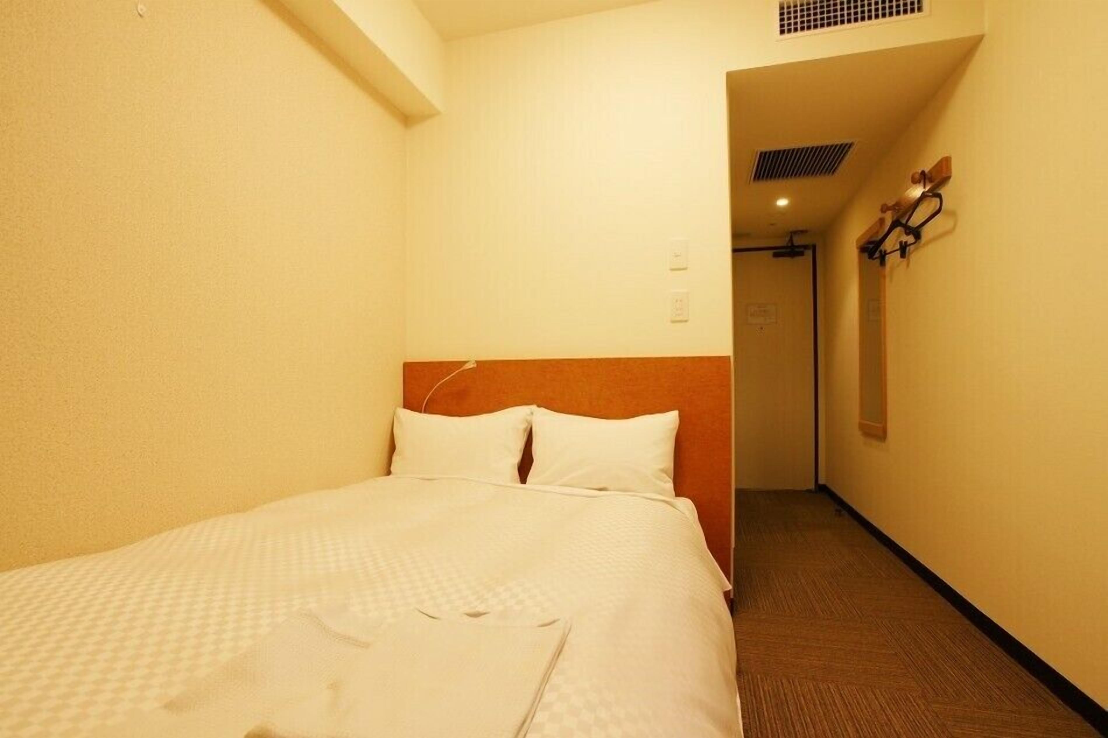 A hotel room with a bed, a wooden headboard, a wall-mounted light fixture, and a door leading to another room.
