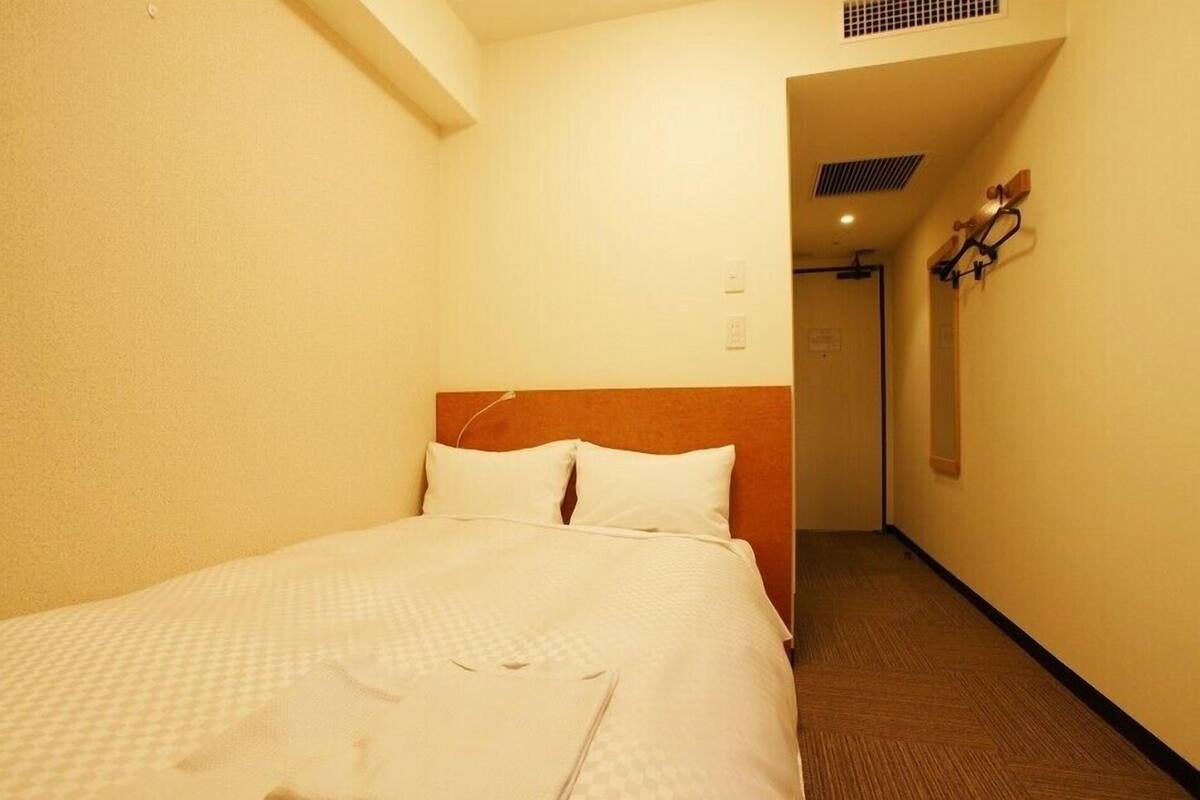 A hotel room with a bed, a wooden headboard, a wall-mounted light fixture, and a door leading to another room.