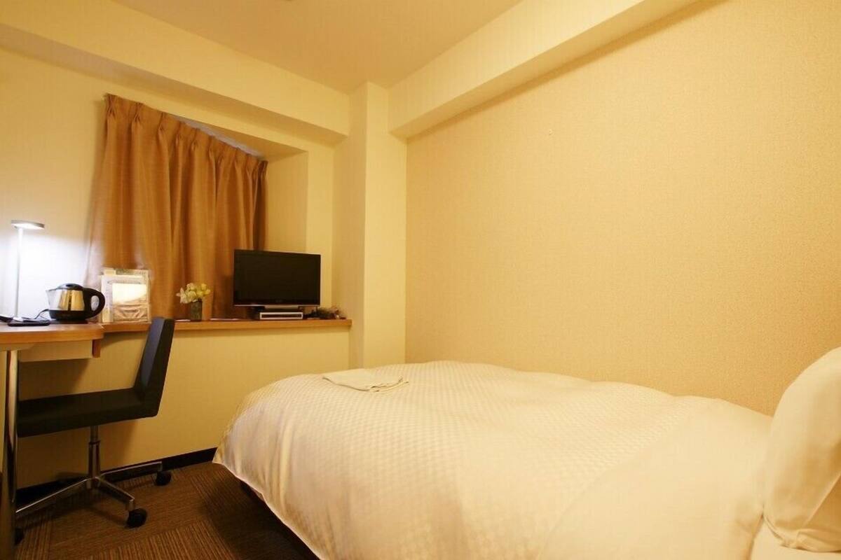A hotel room with a bed, a desk with a computer, a chair, and a window with curtains.