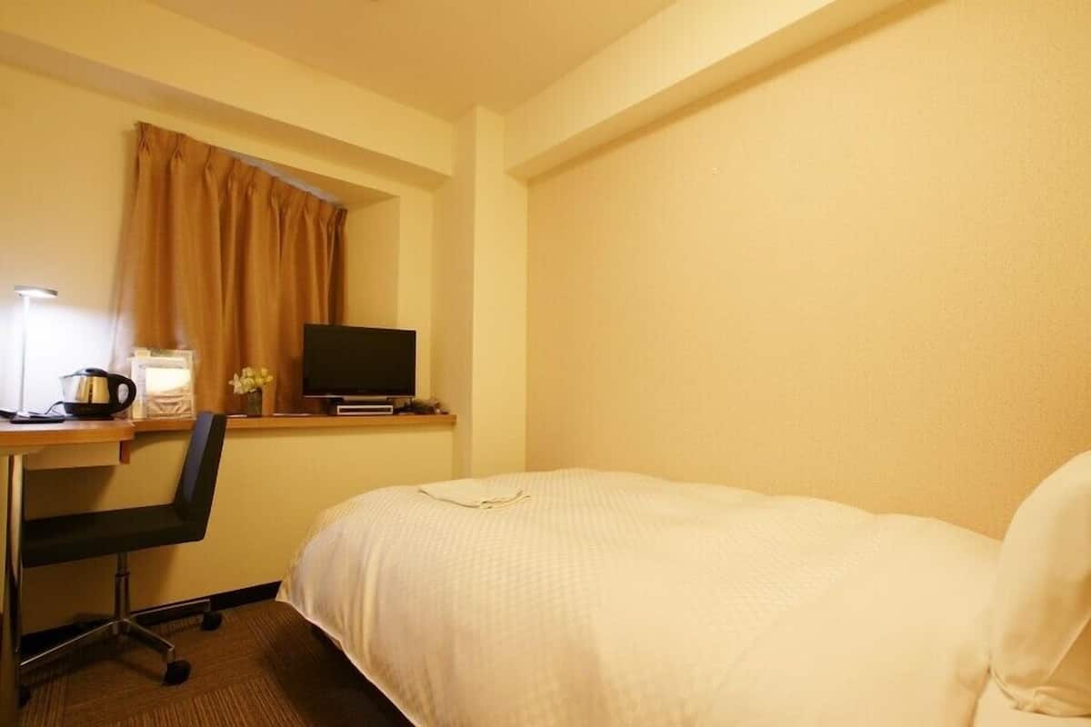 A hotel room with a bed, a desk with a computer, a chair, and a window with curtains.