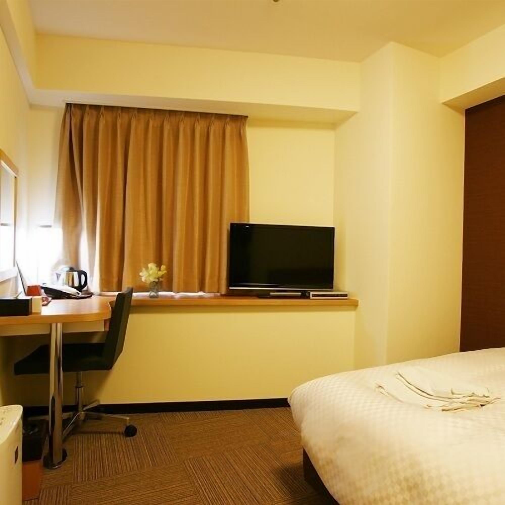 A hotel room with a bed, a desk with a television, a chair, a window with curtains, and a small table with a flower vase.