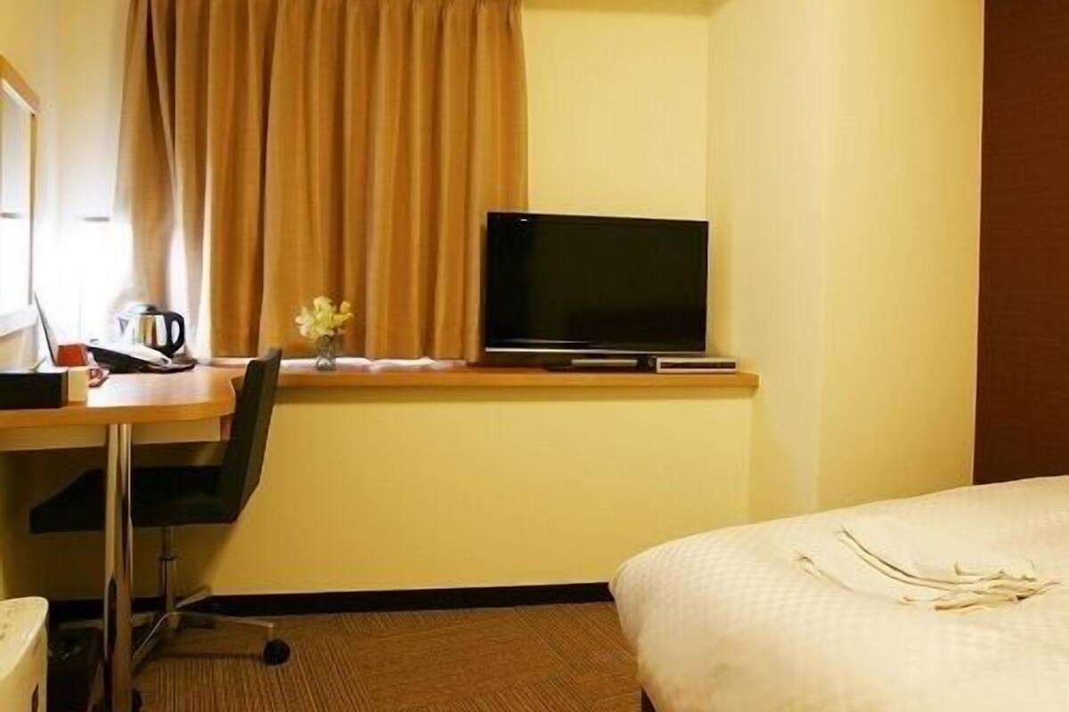 A hotel room with a bed, a desk with a television, a chair, a window with curtains, and a small table with a flower vase.