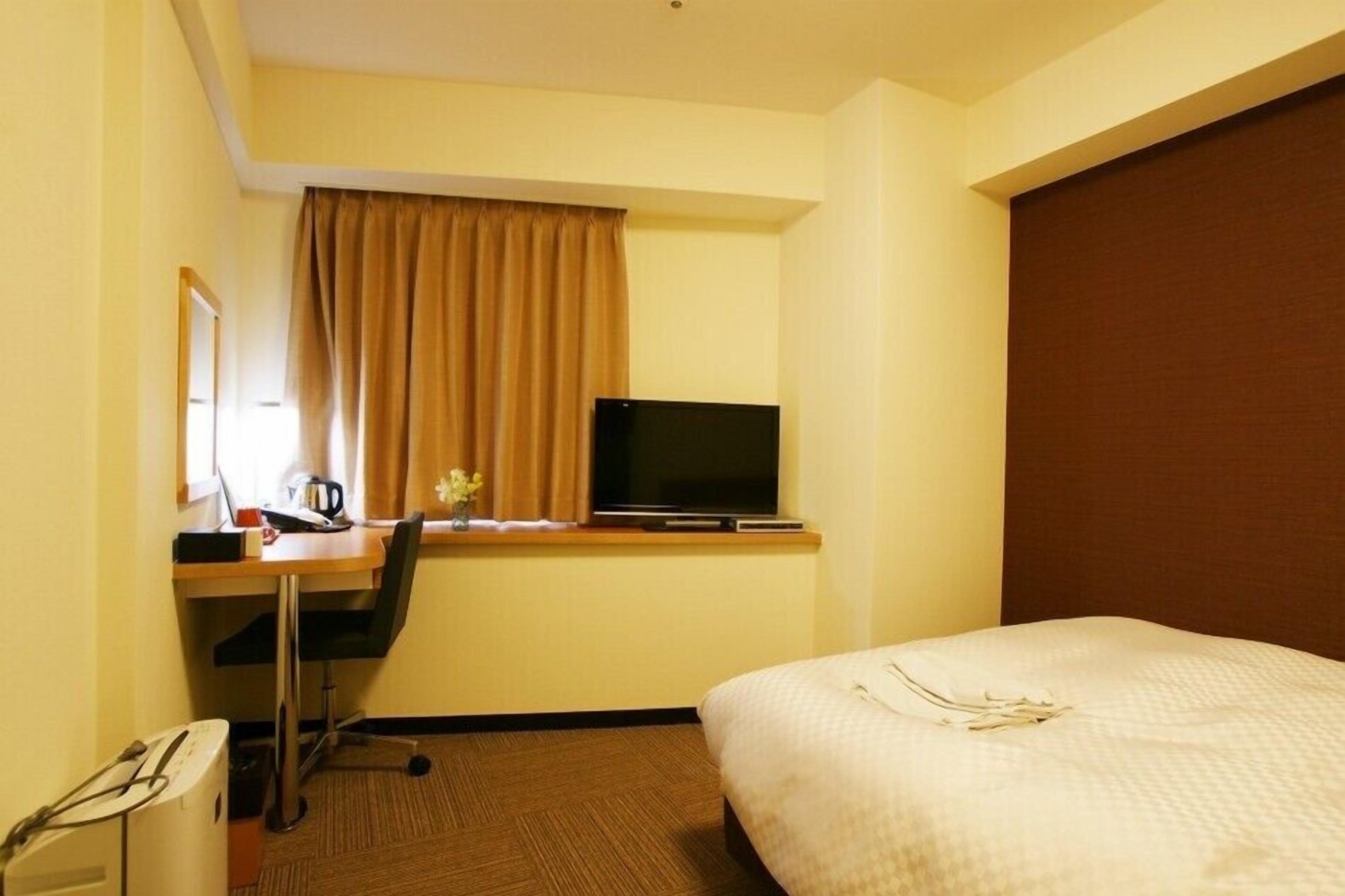 A hotel room with a bed, a desk with a television, a chair, and a small table with a cup and flowers.