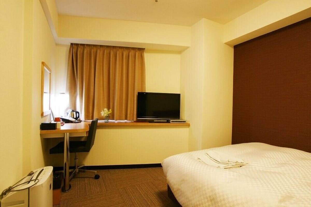 A hotel room with a bed, a desk with a television, a chair, and a small table with a cup and flowers.