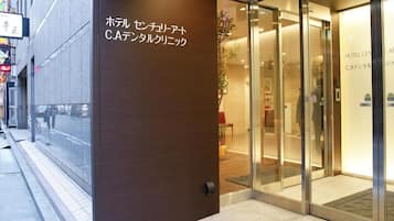 A hotel entrance with a glass door, a sign indicating the hotel name, and a visible reception area inside.