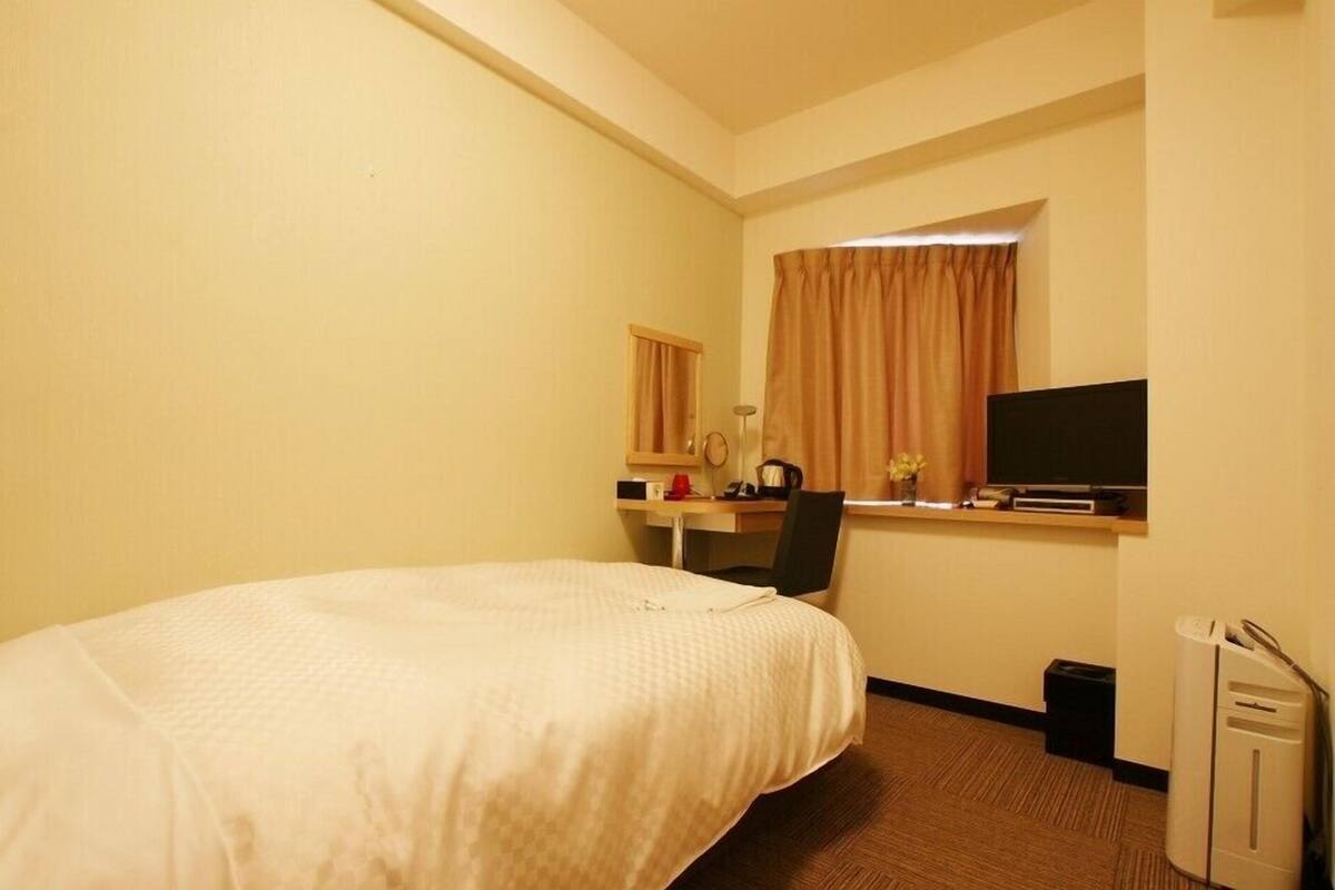 A hotel room with a bed, a desk with a computer, a TV, and a window with curtains.