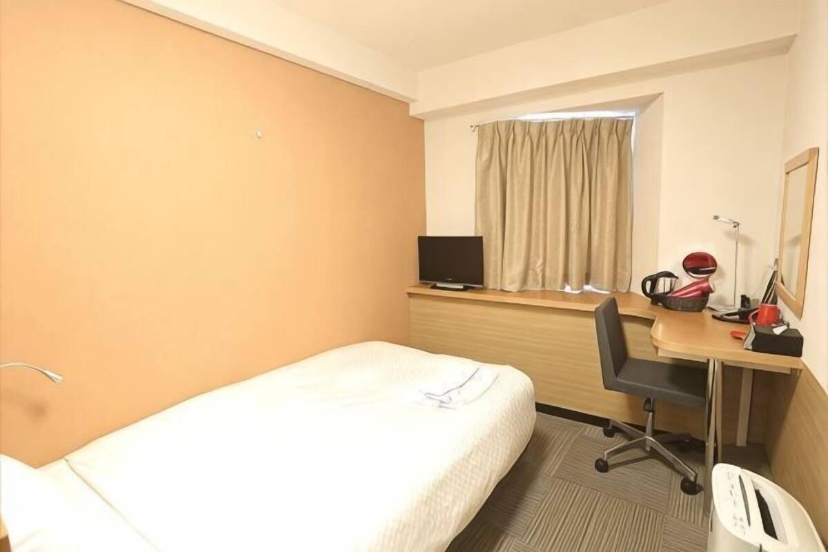 A hotel room with a single bed, a desk with a chair, a television, and a window with curtains.