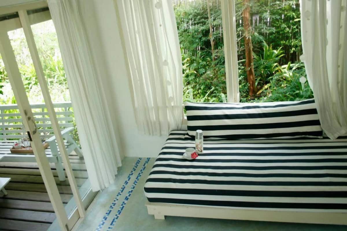 Cama com roupa de cama listrada em preto e branco, mesa de cabeceira com uma xícara e um termo, e vista para vegetação exuberante através de janelas amplas.