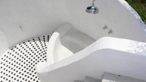 Una bañera exterior blanca con un cabezal de ducha metálico, un desagüe y un escalón pequeño para entrar.