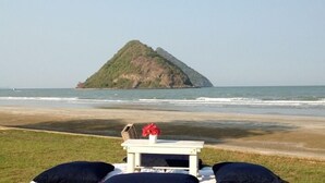 Vista de playa con una pequeña isla en la distancia, una mesa blanca con una flor roja y almohadones negros sobre la arena.
