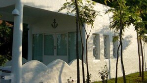 Un edificio blanco con techo plano y ventanas amplias, rodeado de árboles y una montaña al fondo.