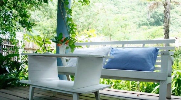 Un espacio exterior con mobiliario blanco, que incluye una banca y una pequeña mesa, rodeado de abundante vegetación y una terraza de madera.