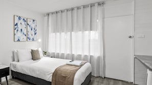 Habitación de hotel con cama, mesita de noche, lámpara de pared y ventana con cortinas traslúcidas.