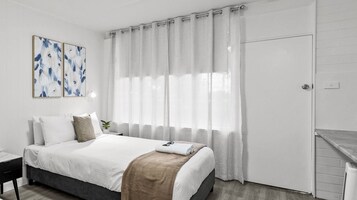 Habitación de hotel con cama, mesita de noche, lámpara de pared y ventana con cortinas traslúcidas.