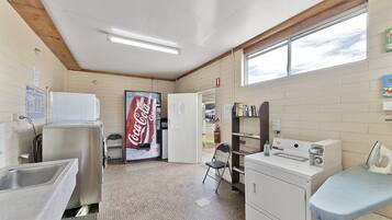 Una sala de lavandería con lavabos, refrigerador, tabla de planchar y máquina expendedora de Coca-Cola.