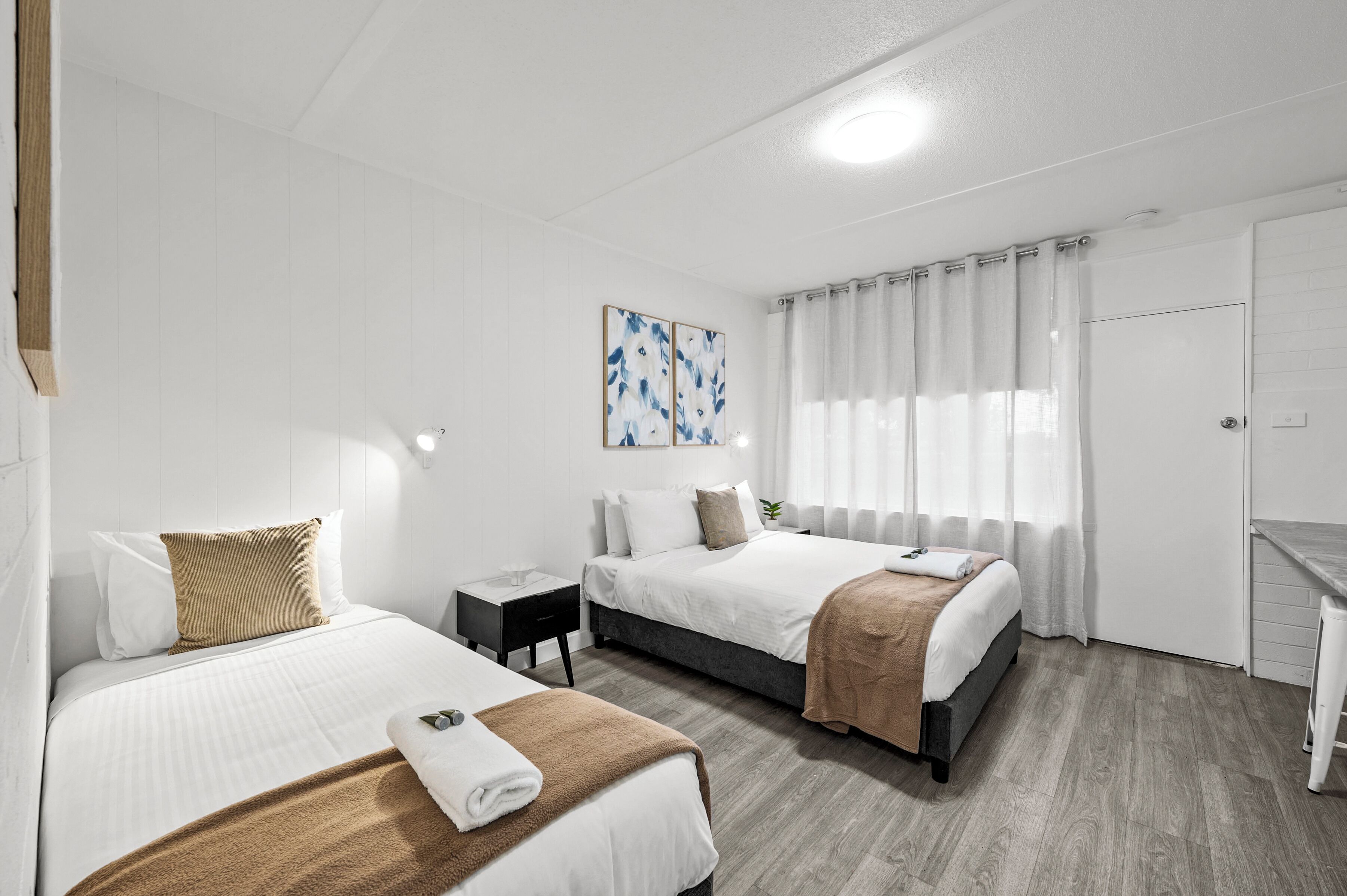 A hotel room with two beds, a nightstand, a painting, and a window with curtains.
