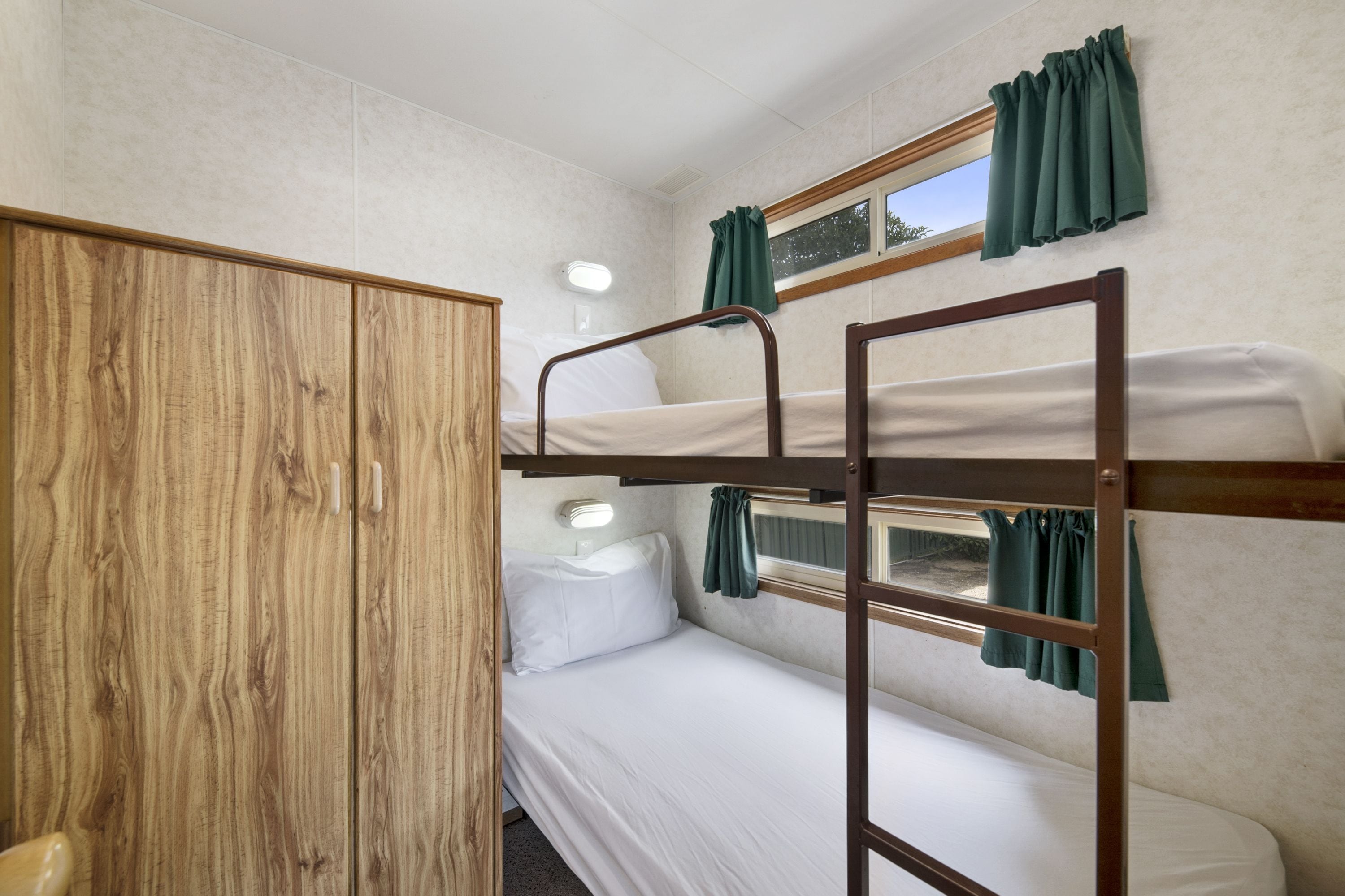 A bunk bed room with a wooden wardrobe, a bedside table, and a window with curtains.