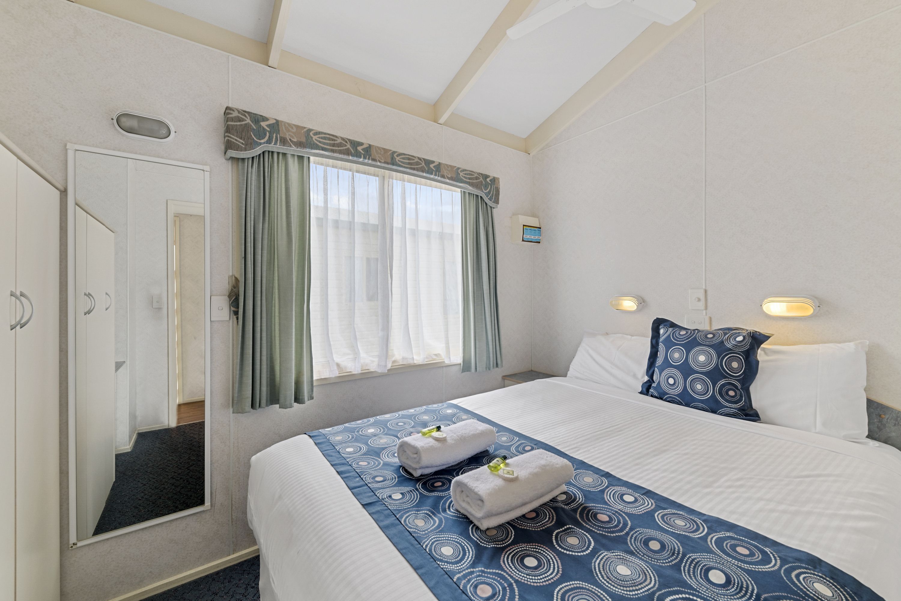 A hotel room with a bed, a mirror, a window with curtains, and a wall-mounted light fixture.