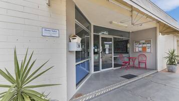 L’entrée d’un immeuble, avec une pancarte indiquant « Vente de logements », une porte vitrée et un petit espace extérieur aménagé avec des chaises rouges.