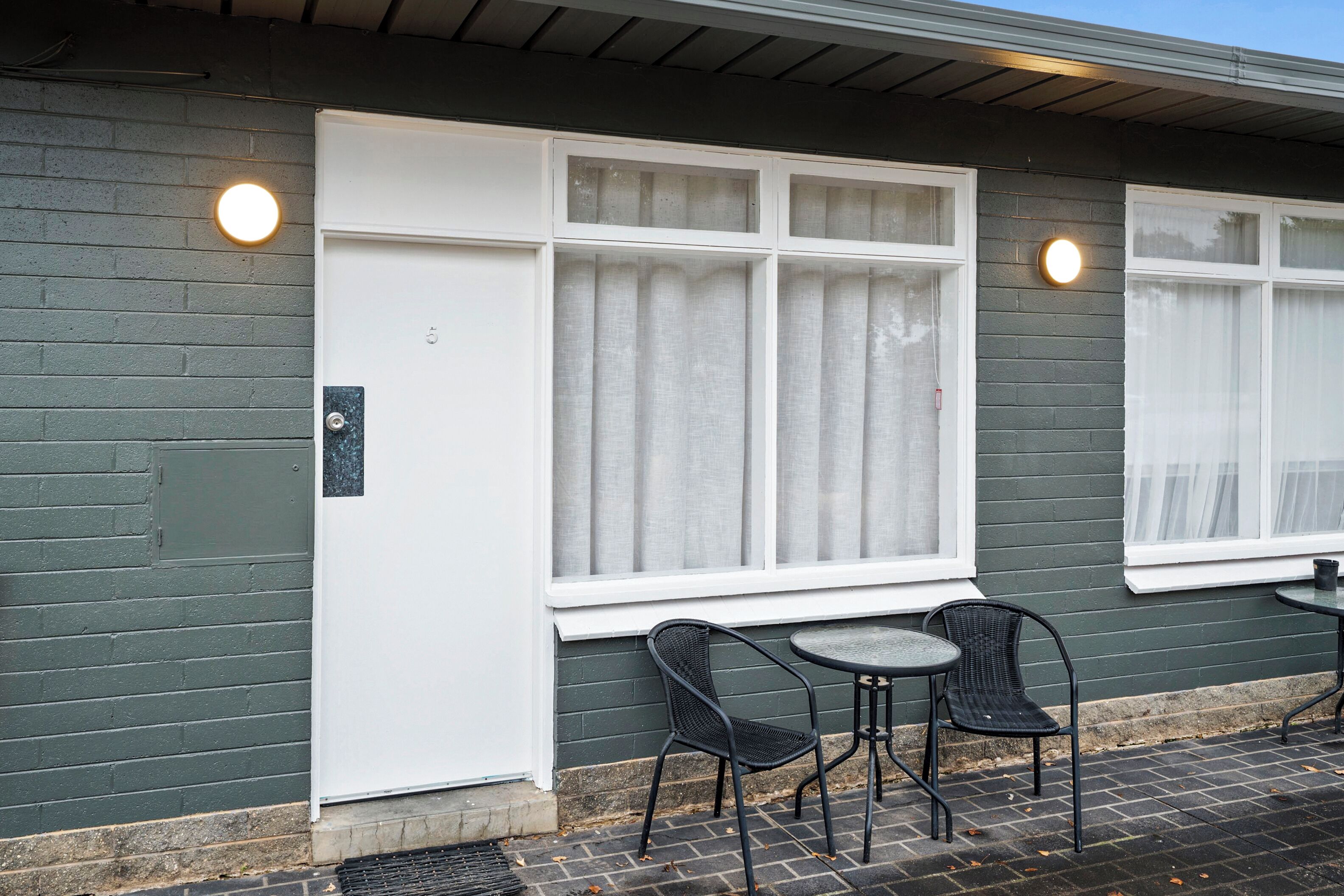 Um edifício com uma porta branca, duas janelas com cortinas e duas mesas de exterior com cadeiras.