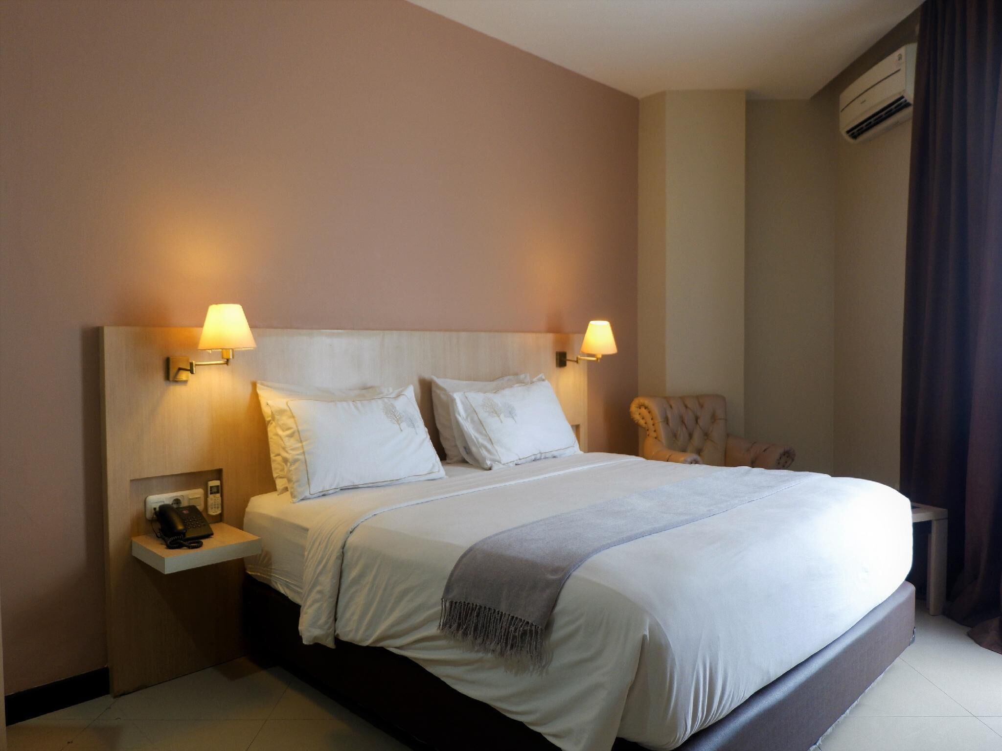 A hotel room with a large bed, two bedside lamps, a telephone, and an air conditioning unit.